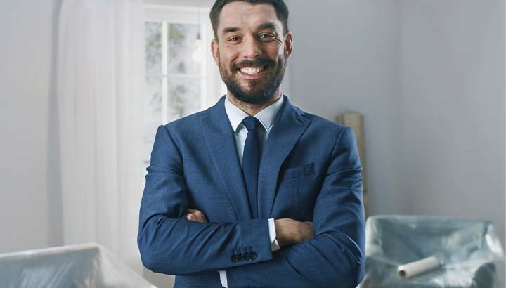 Fotografia de um corretor de imóveis homem, com braços cruzados, vestindo terno azul.