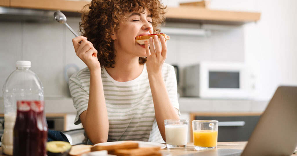 Uma mulher jovem mordendo uma torrada na cozinha. Ela está apoiada em uma mesa, onde há copos com bebidas e outras comidas.