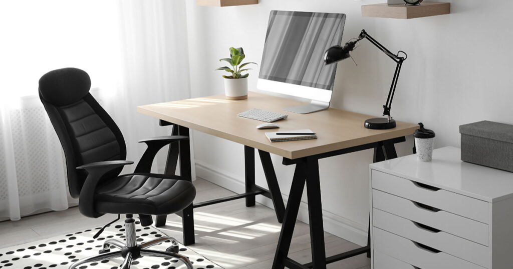 Móveis para home office em um quarto pequeno de uma casa. Há uma mesa com um computador, bloco de anotações e um vaso de planta, assim como uma cadeira.