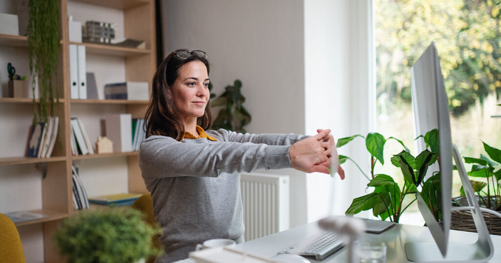 Mulher se alongando em um escritório em casa. Há estantes com livros e plantas no fundo, uma mesa com um computador e vasos de plantas e grandes janelas.